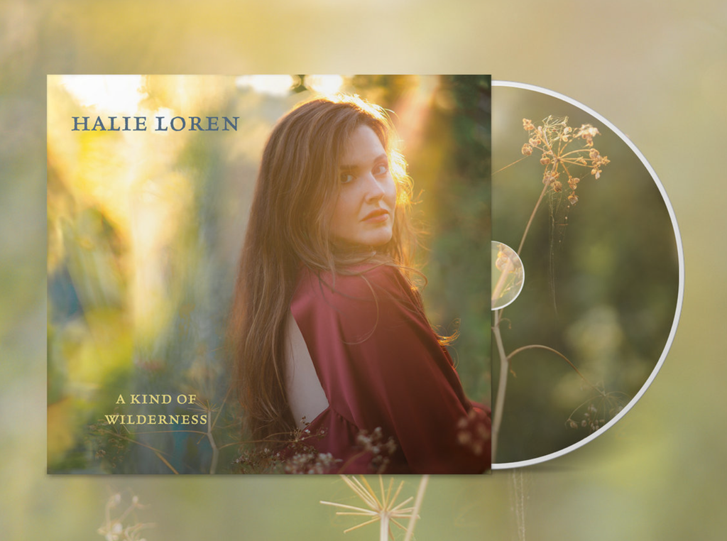 Album cover of Halie Loren's 2026 album "A Kind of Wilderness" picturing Halie Loren wearing a red shirt, backlit by sunbeams and looking over her right shoulder against a background of watercolor-like textures in shades of blue, green, and yellow. To the right of this image is half of a Compact Disc featuring an image of an umbillifer plant that has gone to seed.